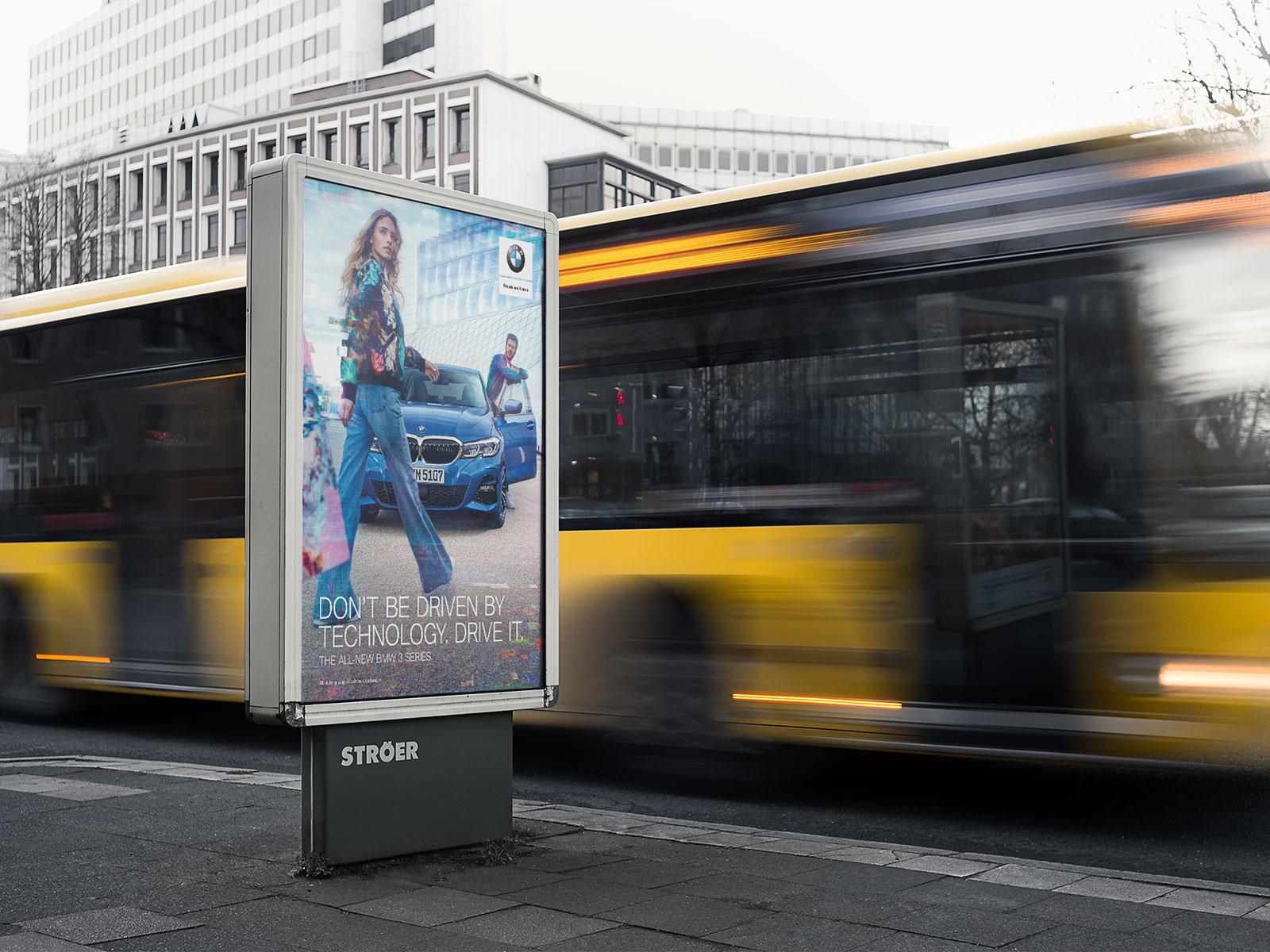 BMW Citylight-Poster an Ströer-Säule in urbaner Straßenszene: BMW 3er mit zwei Personen, Headline „Don't Be Driven By Technology. Drive It."